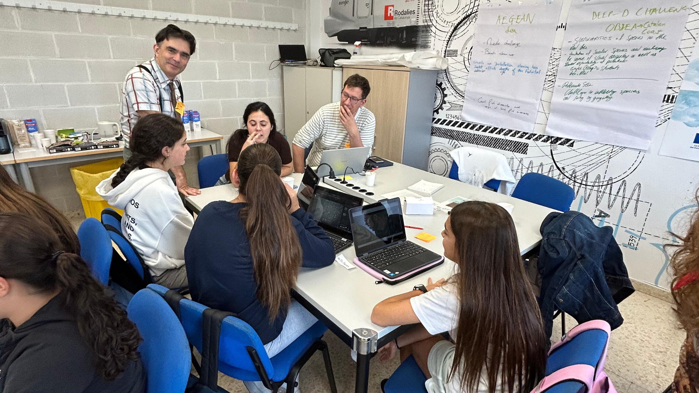 Auf dem Bild sitzt eine Gruppe Sch&uuml;ler*innen mit Laptops um einen Tisch herum. Am Tischende steht eine Lehrperson und erkl&auml;rt etwas, dem die Sch&uuml;ler*innen aufmerksam zuh&ouml;ren.