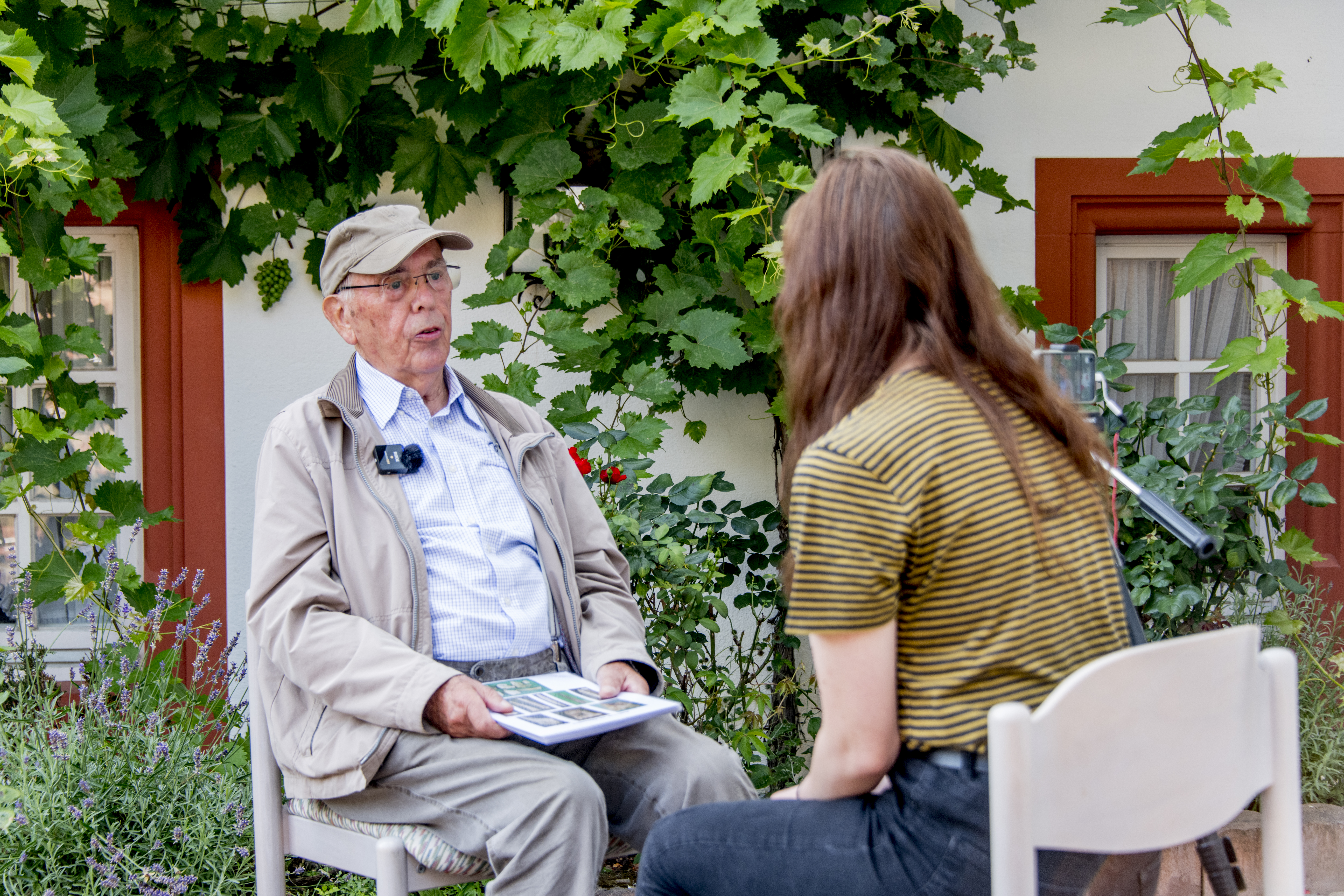 Ein älterer Herr sitzt mit einem Mikro ausgestattet vor einer weinbewachsenen Hauswand und wird von einer Person interviewt.