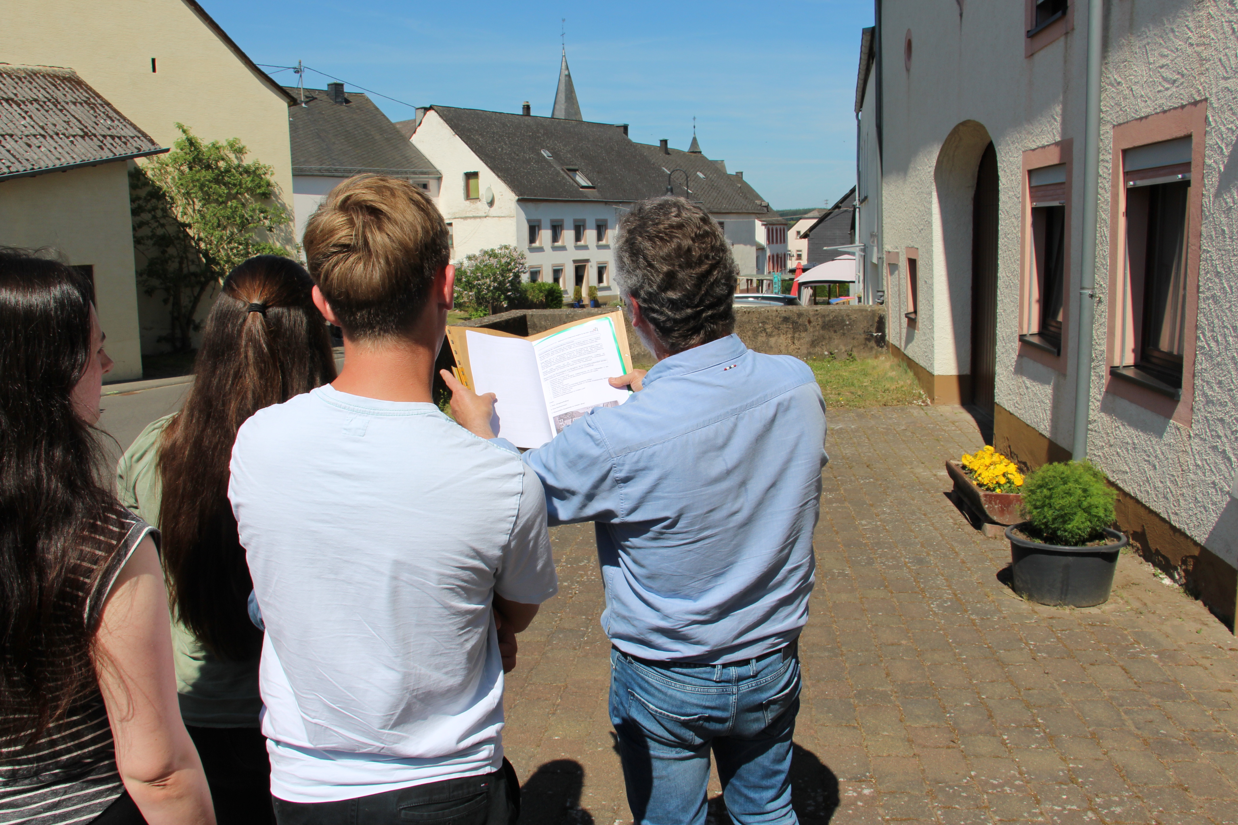 In einer Gasse stehen Menschen mit dem Rücken zum Betrachter:in und gucken gemeinsam ein Dokument an. 