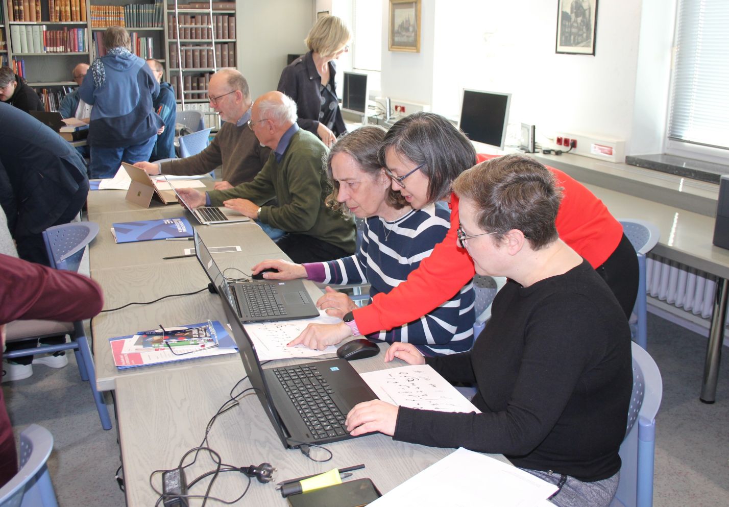 Auf dem Bild sind mehrer Menschen zu sehen die um einen Tisch versammelt vor ihren Laptops sitzen und historische Dokumente transkribieren. 
