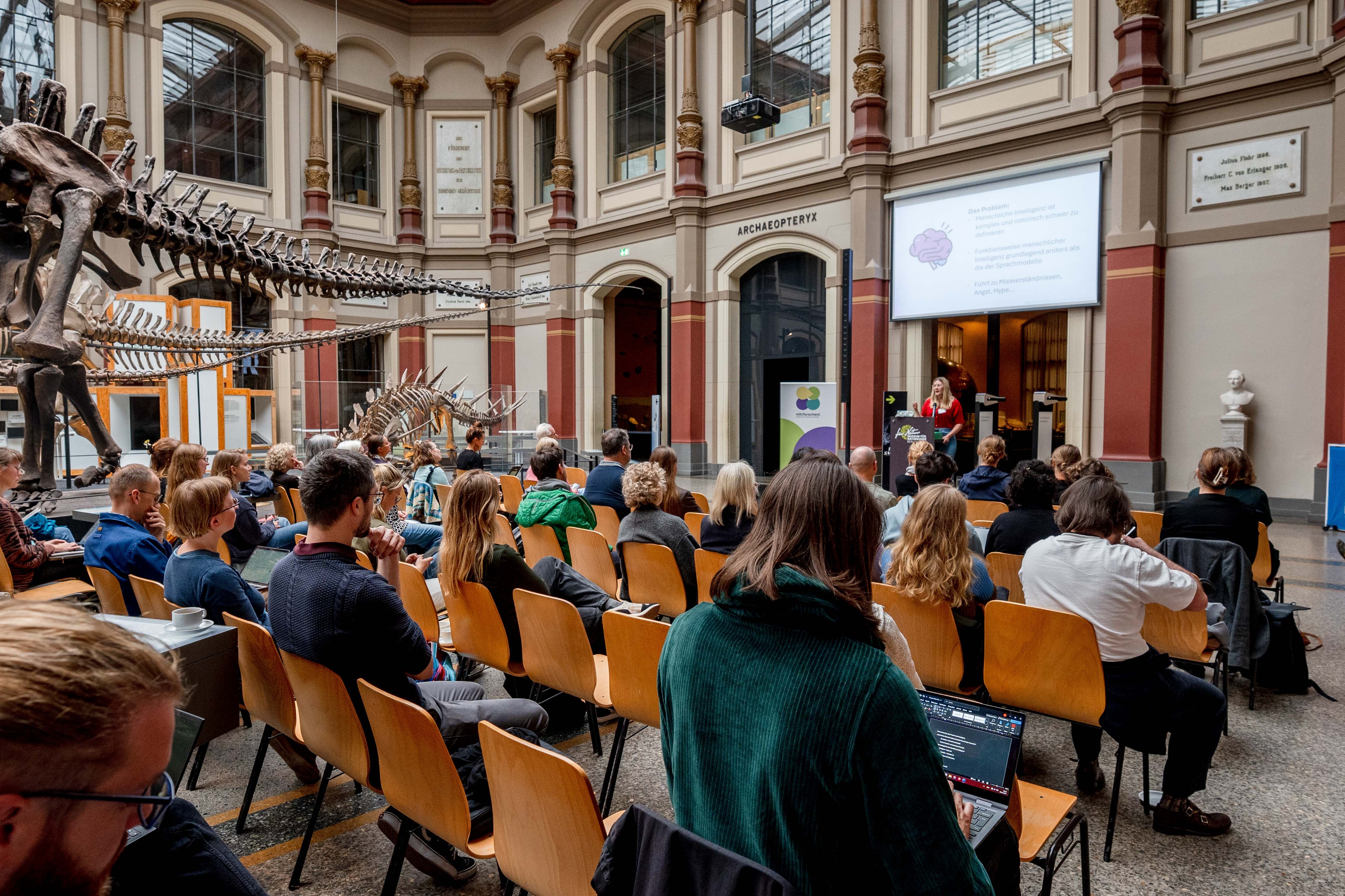 Viele Menschen sitzen in hölzernen Stuhlreihen im Museum für Naturkunde und lauschen der Vortragenden. Im Hintergrund sind die prächtigen Räumlichkeiten und Ausstellungsstücke des Museums zu sehen inklusive des großen Dinosaurierskeletts.