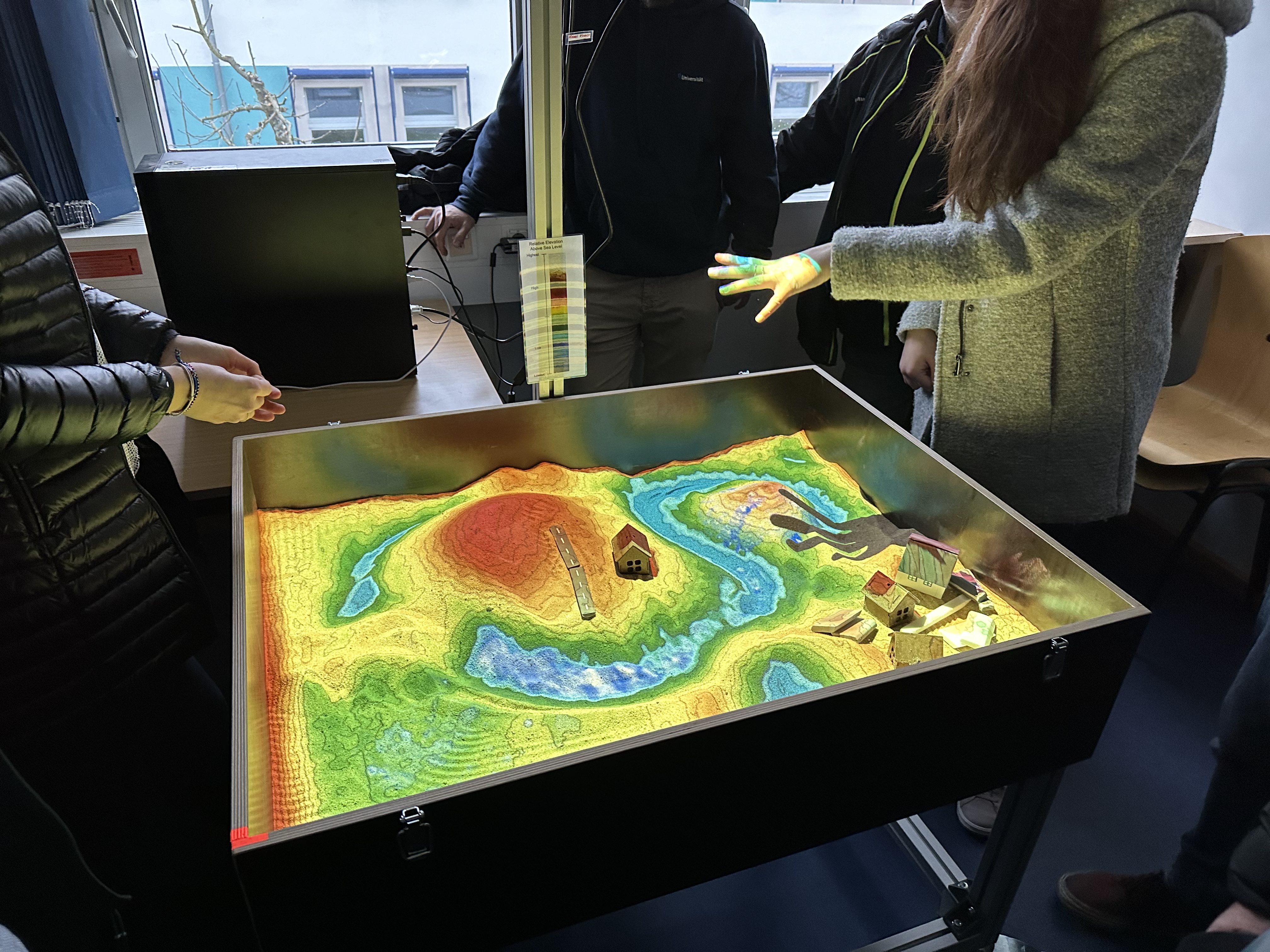 Auf dem Bild ist stehen Menschen um einen Kasten herum, in dem eine Miniatur einer Topographie zu sehen ist. Dafür ist Sand aufgeschüttet, der mit entsprechend ansteigender Höhe in unterschiedlichen Farben angestrahlt ist. An den tiefsten Stellen ist Wasser zusehen. Darüber geht es von grün über gelb bis hin zu rot für die höchsten Stellen. 