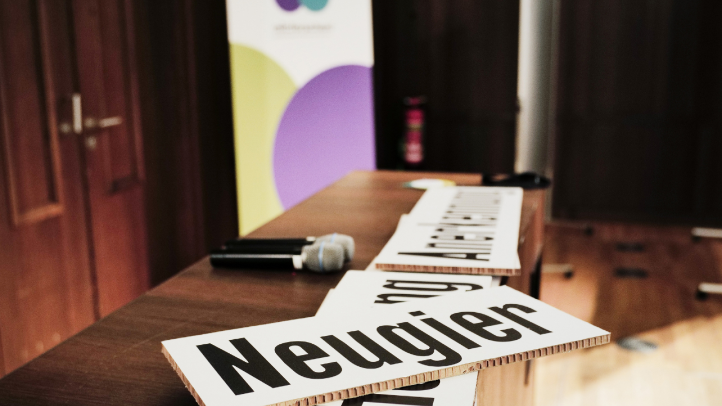 Foto: Jann Wilken / mit:forschen! Ein Schild mit der Aufschrift "Neugier" und zwei Mikrofone liegen auf einer Bühne. Der Hintergrund ist verschwommen.